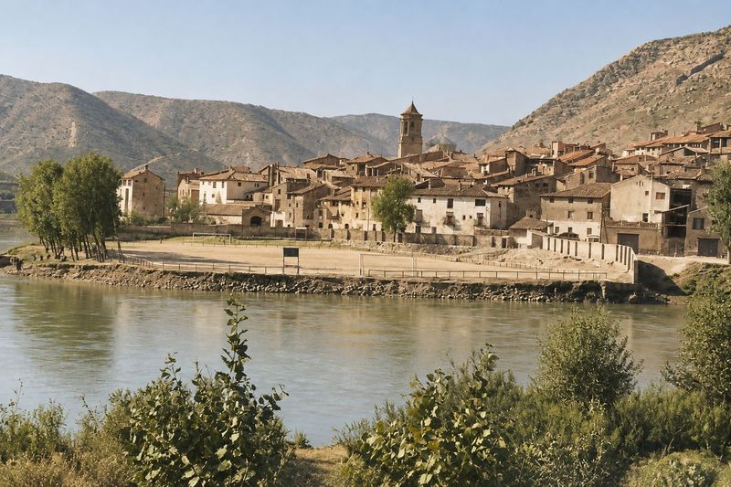 El caserío escalonado y el campanario, con el campo de fútbol y su cercado delante, vistos desde la otra orilla del río, con matorral en primer plano.