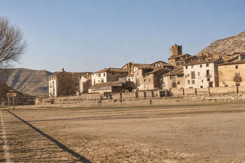 Un campo de tierra con una tapia baja detrás y, sobre ella, casas con balcones y un campanario; a la izquierda, un árbol sin hojas y un cartel junto al campo.