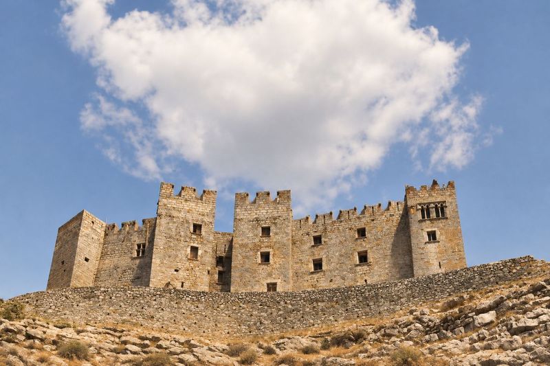 Cuatro torres almenadas del castillo sobre un muro de contención de piedra, con una nube grande detrás; la torre de la derecha tiene una ventana geminada.