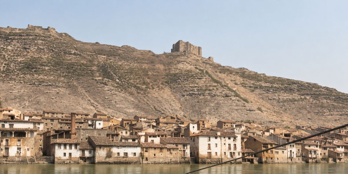 Casas sobre la orilla bajo el cerro, con las ruinas del castillo en lo alto y una muralla que baja de la cima; una chimenea alta sobresale y un cable cruza la imagen.