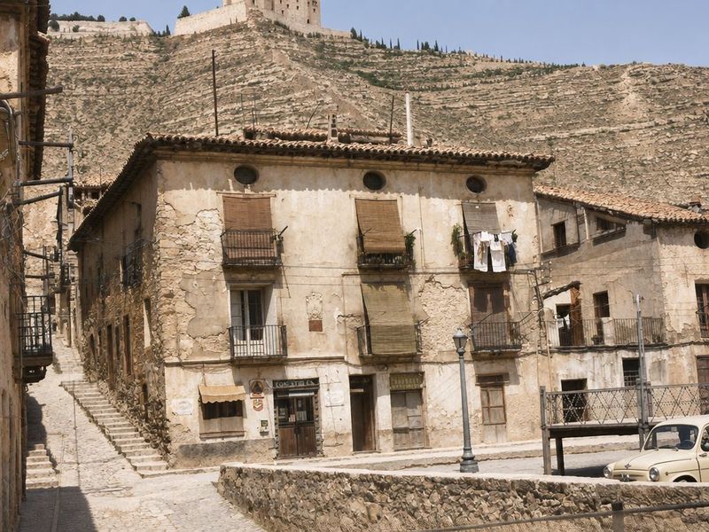Casa de esquina con alero moldurado y óculos, una tienda en la planta baja, farola y coche pequeño, junto a una calle en escalera; sobre el cerro, el castillo.