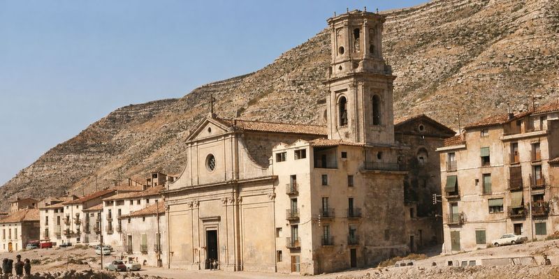 Fachada de la iglesia con su frontón y la torre sin cupulín, un bloque de casas adosado a la derecha y el cerro de estratos detrás.
