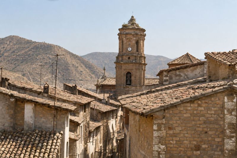 Tejados de teja y casas de ladrillo en una calle estrecha, con la torre de la iglesia, con su nido, y los montes detrás.