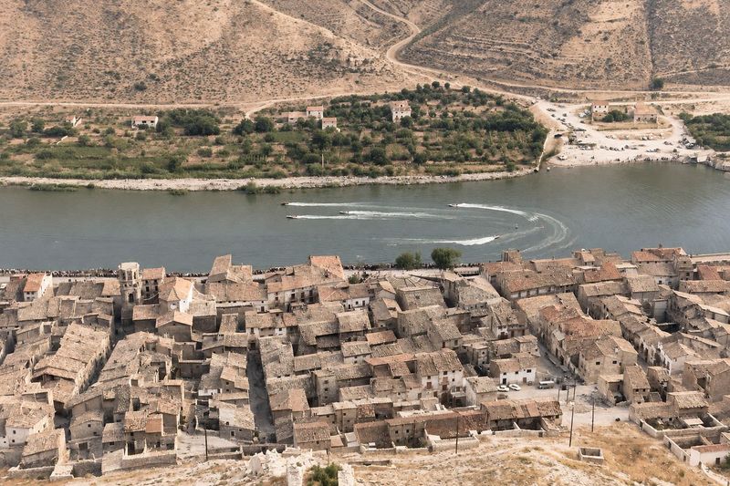 Tejados y torre de la iglesia vistos desde la ladera del castillo; en el río, lanchas con sus estelas en una carrera, y al otro lado una isla de huertas.