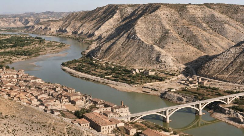 El puente de arcos sobre el Ebro junto a naves, una chimenea y casas del extremo del pueblo; el río se curva entre huertas bajo montes de ladera pelada.
