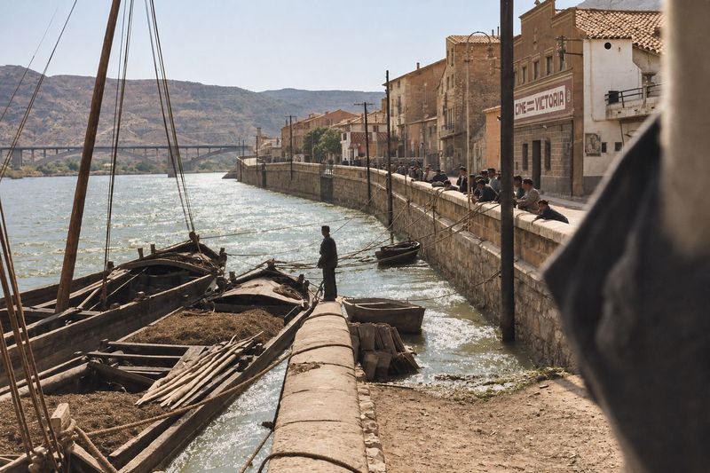 El muro en curva con farolas junto a la fachada del Cine Victoria; llauts amarrados con carga, un hombre de pie, jóvenes en el pretil y el puente al fondo.