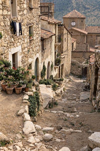Callejón de piedra en escalera, con macetas delante de las casas, ropa tendida y una torre con reloj al fondo.