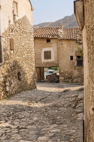Un paso cubierto bajo una casa sobre pilares, al final de una calle empedrada, con un coche pequeño y un carro bajo la casa y muros de mampostería.