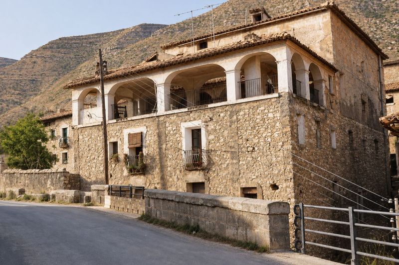 Casa de mampostería de tres plantas con una solana de arcos encalados sobre la carretera, con un pretil y una barandilla delante, y un monte seco detrás.