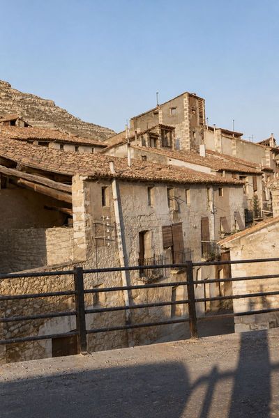 Casas de mampostería de dos a cuatro plantas con tejados de teja, un balcón de forja y postes con aisladores, bajo un cerro y vistas tras una barandilla.