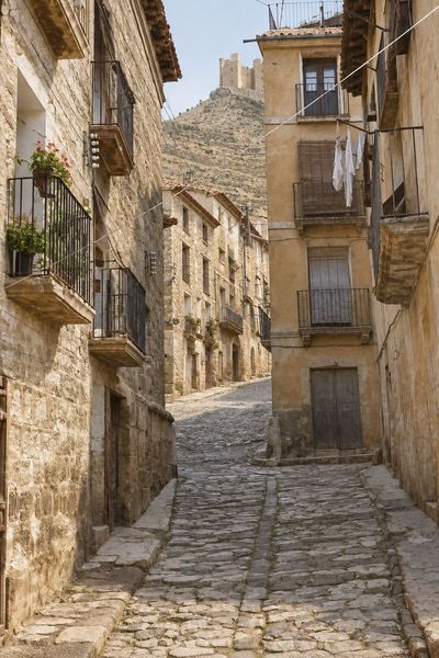 Calle estrecha de cantos rodados con cuneta central entre casas de tres y cuatro plantas con balcones y ropa tendida; al fondo, el cerro con el castillo.