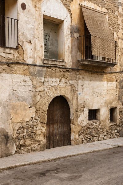 Fachada de mampostería y revoco desconchado con un portal de arco, dos ventanas pequeñas y un balcón con toldo de cañizo, sobre una calle de tierra con acera.
