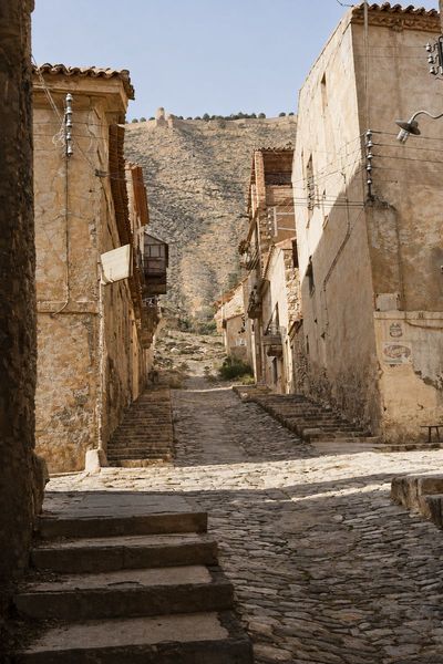 Un cruce de calles empedradas con escalinatas laterales entre casas revocadas, con un farol de brazo y, arriba, la muralla del castillo.
