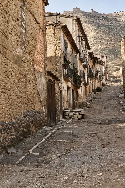 Una calle de tierra en cuesta entre un gran muro de mampostería y una hilera de casas con balcones y un portal de arco; al fondo, el cerro abancalado con la muralla del castillo.