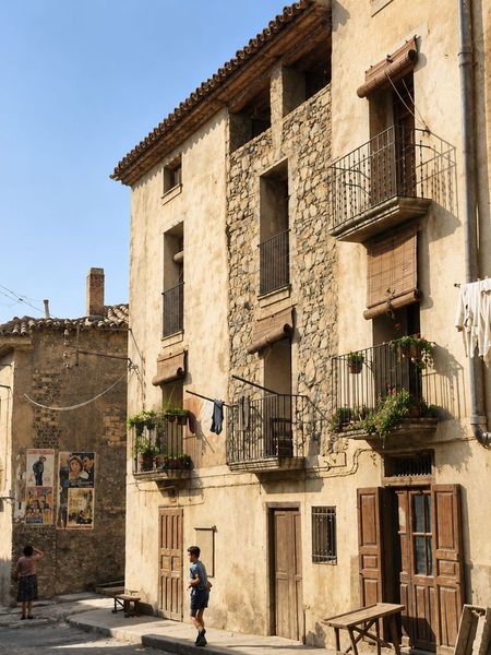 Fachada de cuatro plantas con paño de mampostería, balcones de forja y alero, junto a un muro con carteles de cine; un niño camina por la calle.