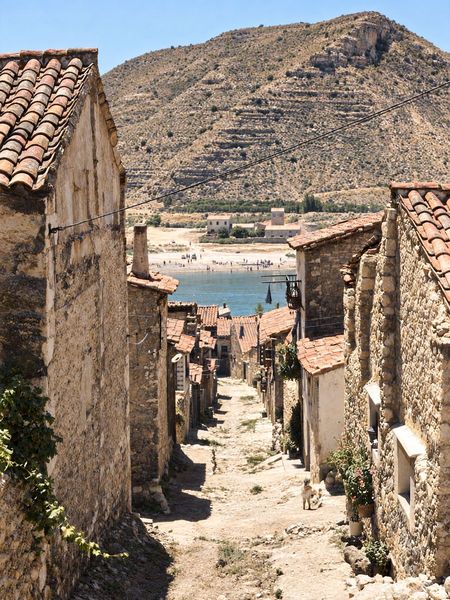Calle estrecha de piedra y tejas que baja al agua, con la playa y el monte de bancales en la otra orilla.