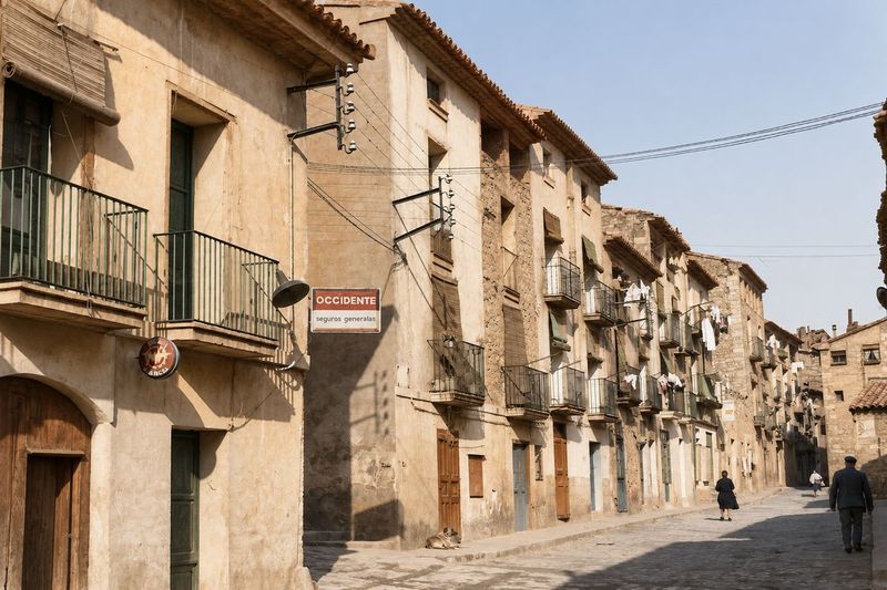 Calle recta con una fila continua de casas de aleros volados y balcones de forja, un letrero de seguros en la esquina y varias personas que caminan.