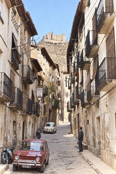 Calle de balcones de forja y persianas enrolladas, con el rótulo de la peluquería, dos Seat de la época y el castillo cerrando la calle.