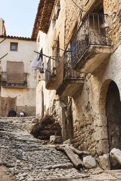 Visto a ras de suelo, un callejón empedrado en cuesta con un portal de arco de sillares, balcones de forja con volutas, leña apilada y unas personas sentadas al fondo.
