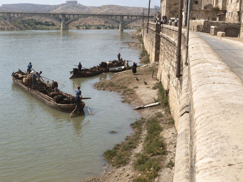 El muro de sillería desciende hasta una playa de grava donde hay llauts cargados con barqueros de pértiga; al fondo se ve el puente y, en lo alto del muro, algunas personas.