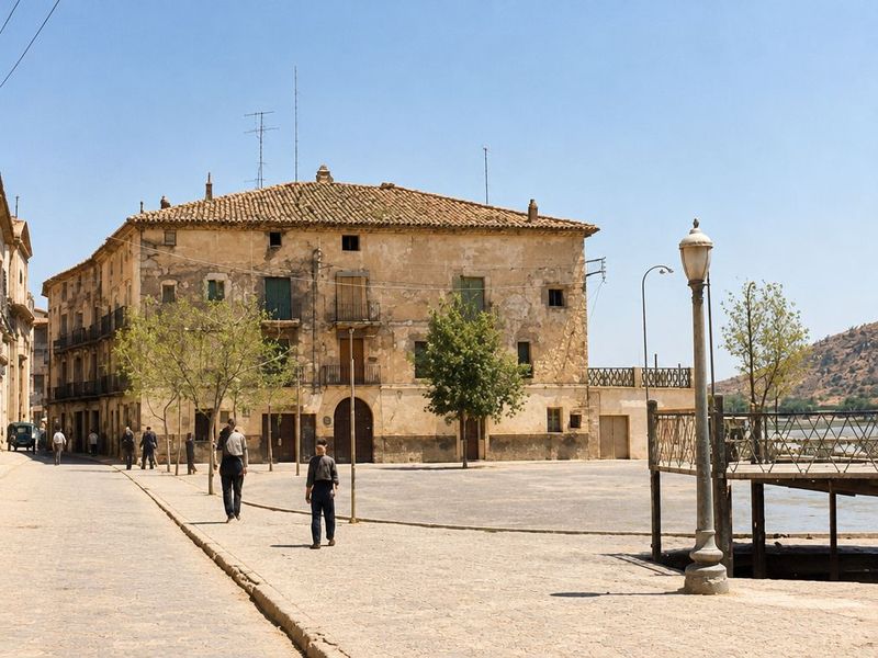 Una plaza amplia con un caserón de portal de arco al fondo, farolas de fundición, árboles jóvenes, un muelle de madera a la derecha y vecinos paseando.