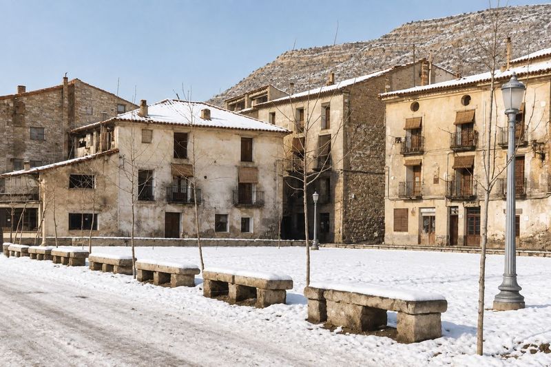 Una plaza cubierta de nieve con bancos de piedra, farolas y árboles jóvenes sin hojas, frente a una hilera de casas bajo la ladera.