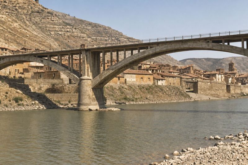 Bajo el arco principal del puente se ven el muro de ribera, las casas, unas barcas y el campanario; en primer plano, la orilla de grava y el río.