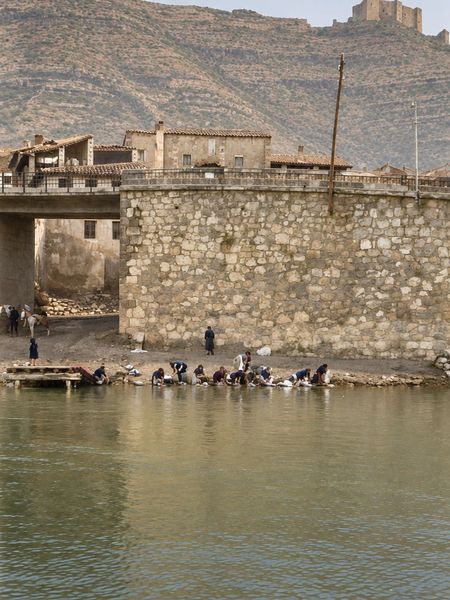 Un muro de mampostería junto a la cabeza de un puente, con casas encima y el castillo en lo alto del monte; en la orilla, un grupo de lavanderas.
