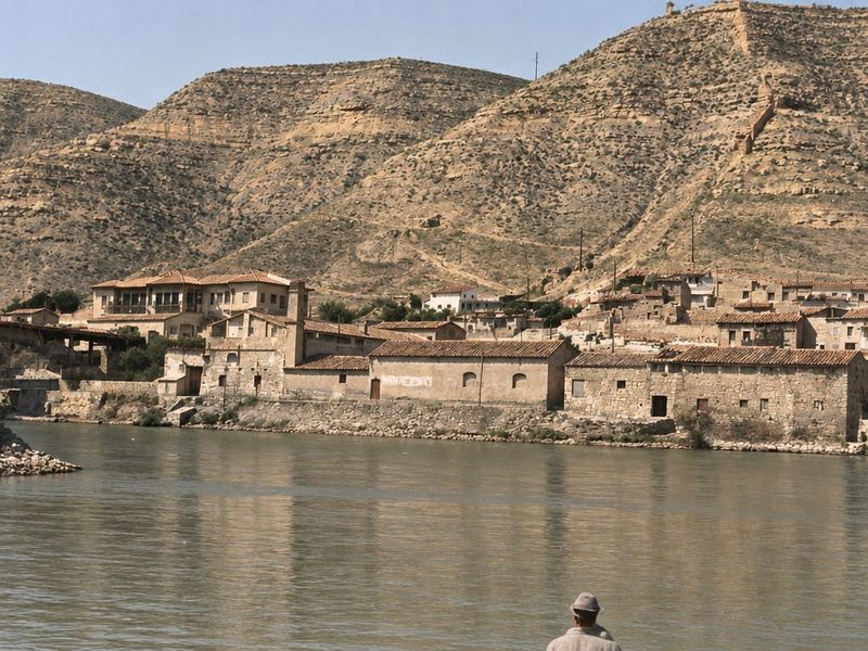 Casas y naves al borde del agua bajo el cerro, con un edificio de galería y torre a la izquierda y una nave de ventanas en arco; abajo asoma un hombre con sombrero.