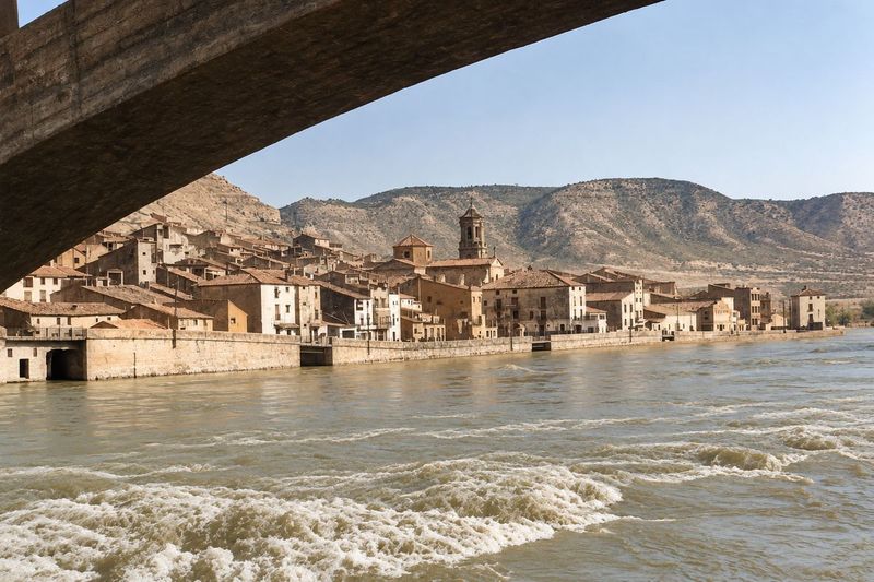 Bajo la viga curva de un puente se ve el frente de casas junto al muro, con la iglesia de cimborrio y torre y una casa alta al final; delante, el río agitado.