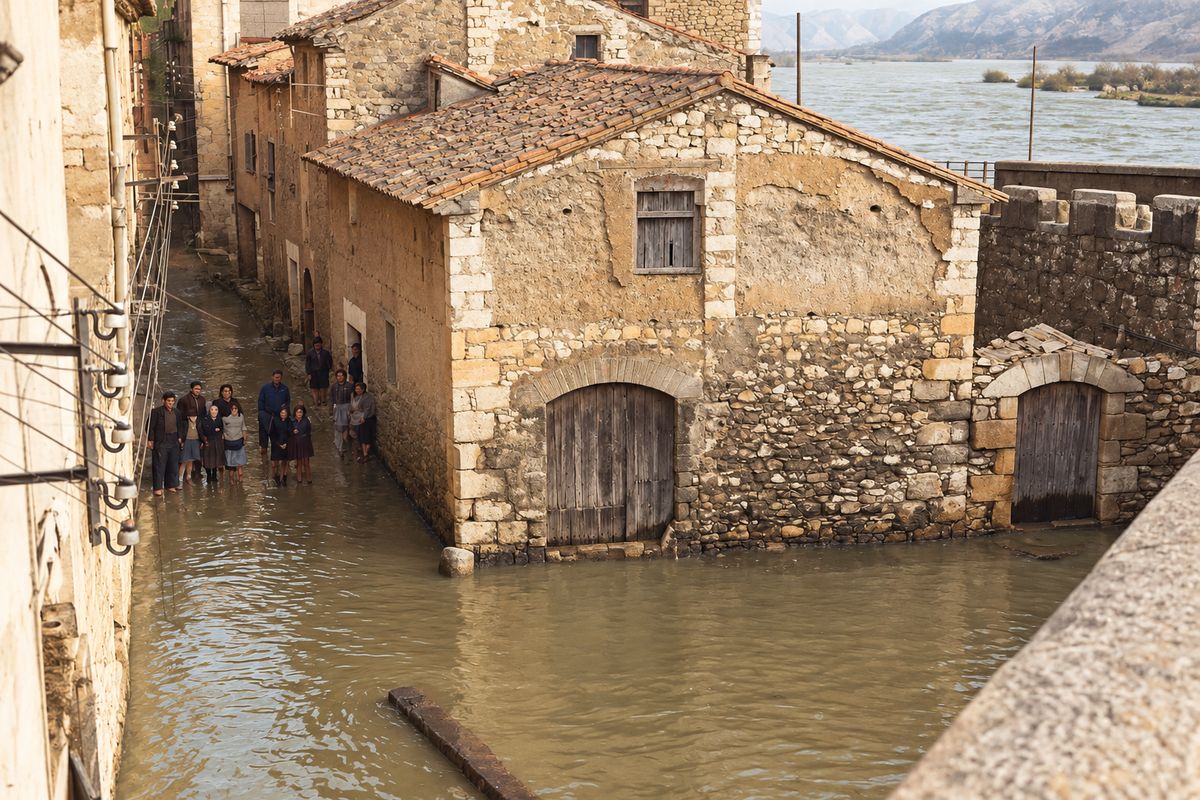 Una casa de piedra con portalón de dovelas, un muro almenado y un portal con tejadillo, con la calle inundada y el río al fondo; a la izquierda, un grupo de personas en la calle.