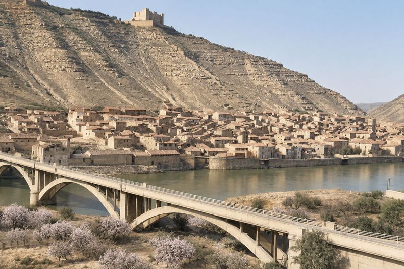 Puente con barandilla en primer plano, el río, el muro de ribera y el caserío escalonado bajo el castillo, con almendros en flor delante.