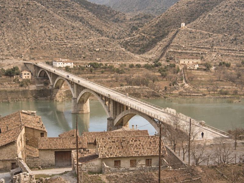 El puente de arcos de hormigón en diagonal sobre el río, con peatones en el tablero y, en primer plano, los tejados de las casas de entrada.