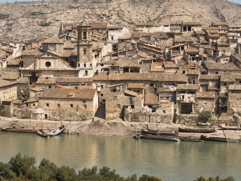 Una orilla de tierra con llauts varados bajo el caserío; la iglesia con óculo y el campanario asoman entre casas escalonadas, con el cerro al fondo.