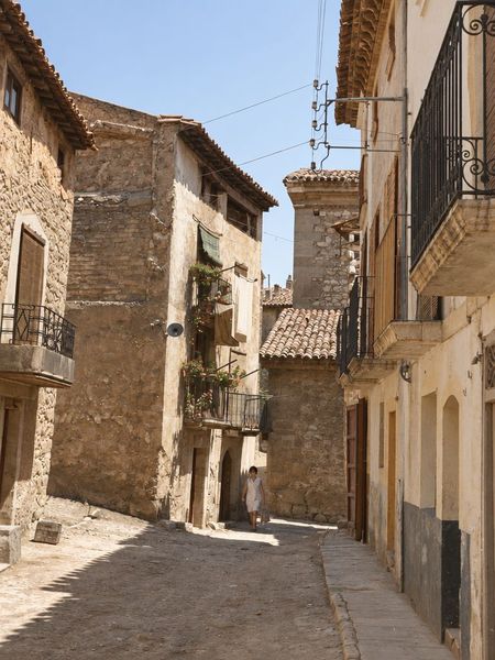 Calle en cuesta entre muros de mampostería y balcones de forja, con sombras marcadas, tejados al fondo y una mujer que camina lejos.