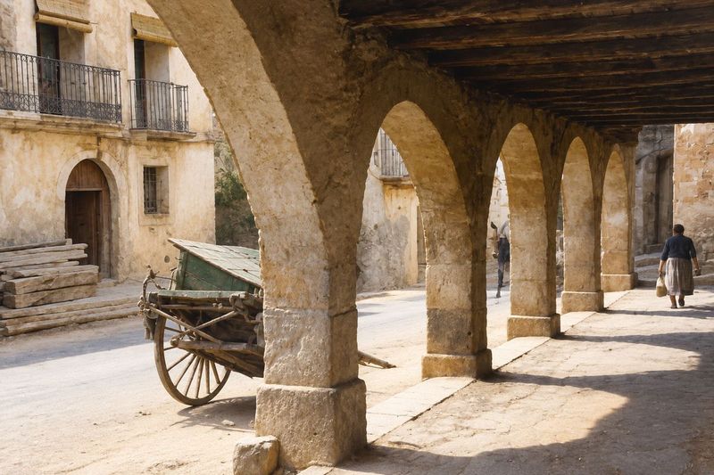 Una galería de arcos de piedra en fuga, con un carro y unos maderos junto a una casa con balcones, y una mujer que camina al fondo.