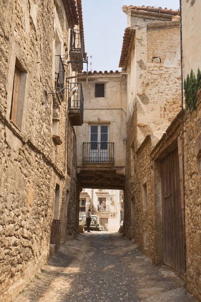 Una calle estrecha entre muros de piedra, con un pasadizo cubierto bajo una casa y, al fondo, una plaza soleada con un coche y una persona.