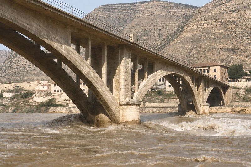 El puente visto desde abajo con el río crecido batiendo la pila, y las casas de la ladera al fondo.