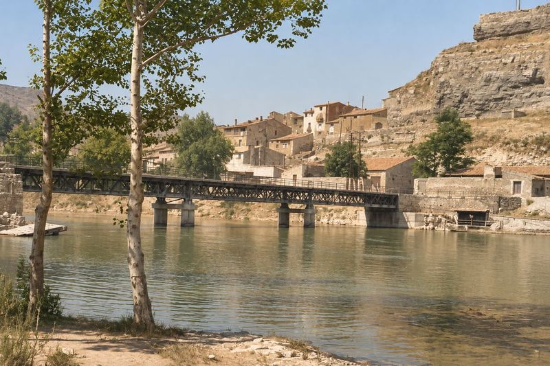 El puente de celosía sobre pilas bajas, con las casas escalonadas bajo el peñasco, un edificio junto al estribo y dos álamos en primer plano.