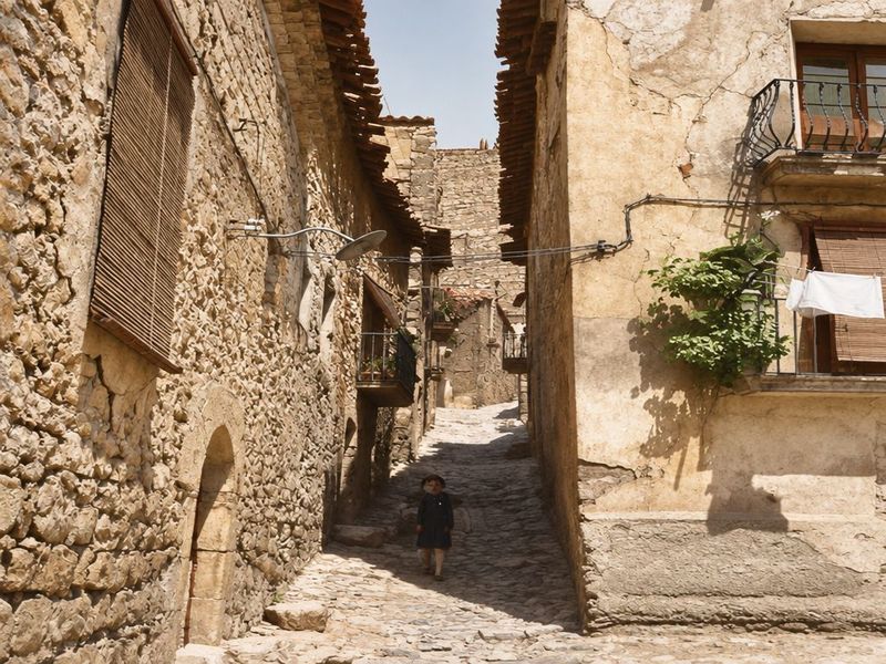 Callejón empedrado entre muros de mampostería, con un portal de arco, un balcón con plantas, ropa tendida, una farola de brazo y una figura al fondo.