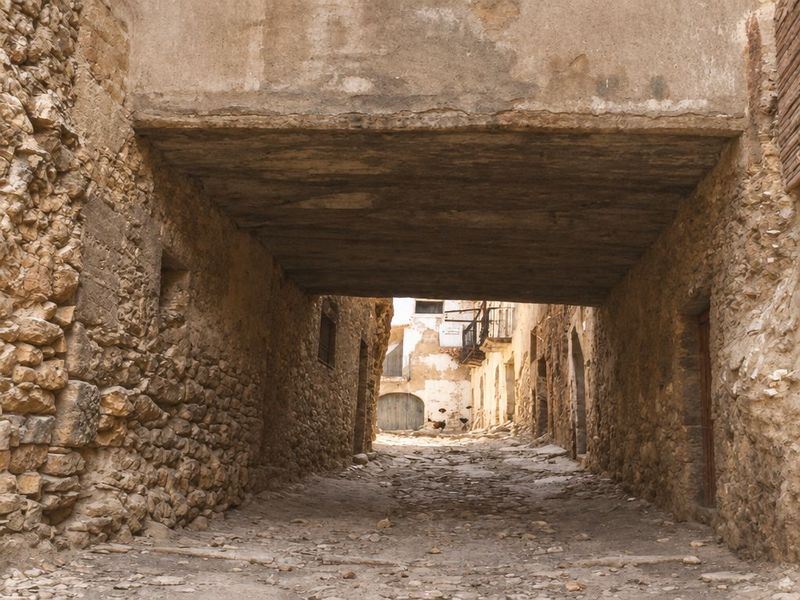 Pasadizo de piedra y vigas sobre una calle empedrada que se abre al fondo a una plaza luminosa, con un balcón y una puerta de arco.