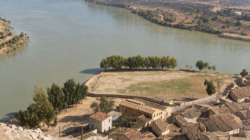 Desde el cerro: el borde del pueblo con tejados y una nave de cubiertas paralelas, y el campo de fútbol con una arboleda junto al río.