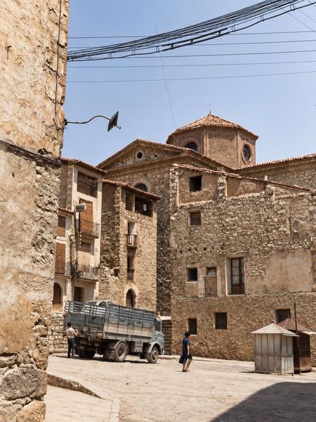 Una plaza de tierra con un camión y dos kioscos de madera, un portal en arco y, al fondo, la iglesia con su óculo y el cimborrio.