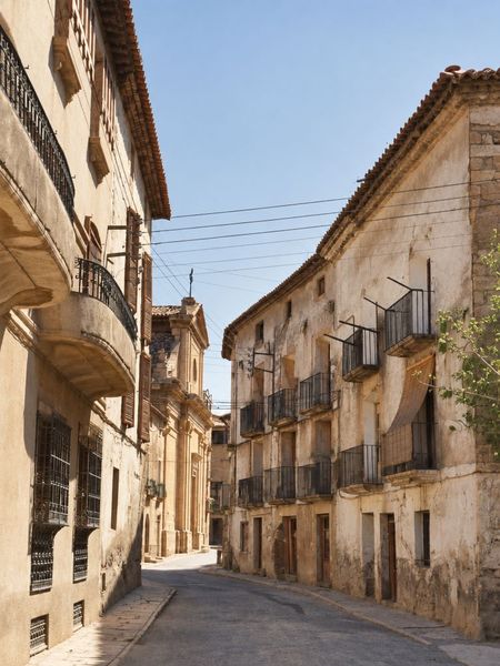 Calle en ligera curva con balcones de forja y aleros a ambos lados; al fondo, un edificio con frontón y un remate metálico sobre la cubierta.