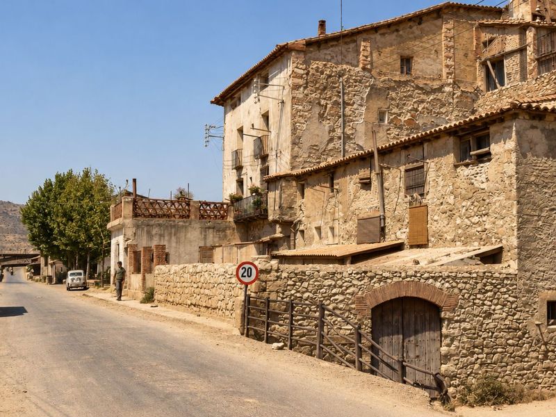 La carretera pasa junto a un muro de piedra con portal de ladrillo y una señal con un 20; detrás, casas desconchadas con galerías y balcones, y un peatón y un coche a lo lejos.