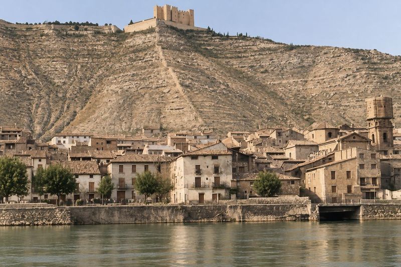 Caserío bajo la ladera abancalada con el castillo en lo alto, un campanario con andamio sobre el muro de ribera y el río en primer plano.