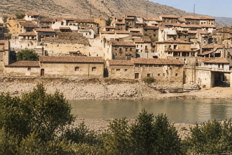 Caserío escalonado sobre naves y un muro junto al río, con galerías y balcones; en primer plano, arbustos y la orilla de grava.